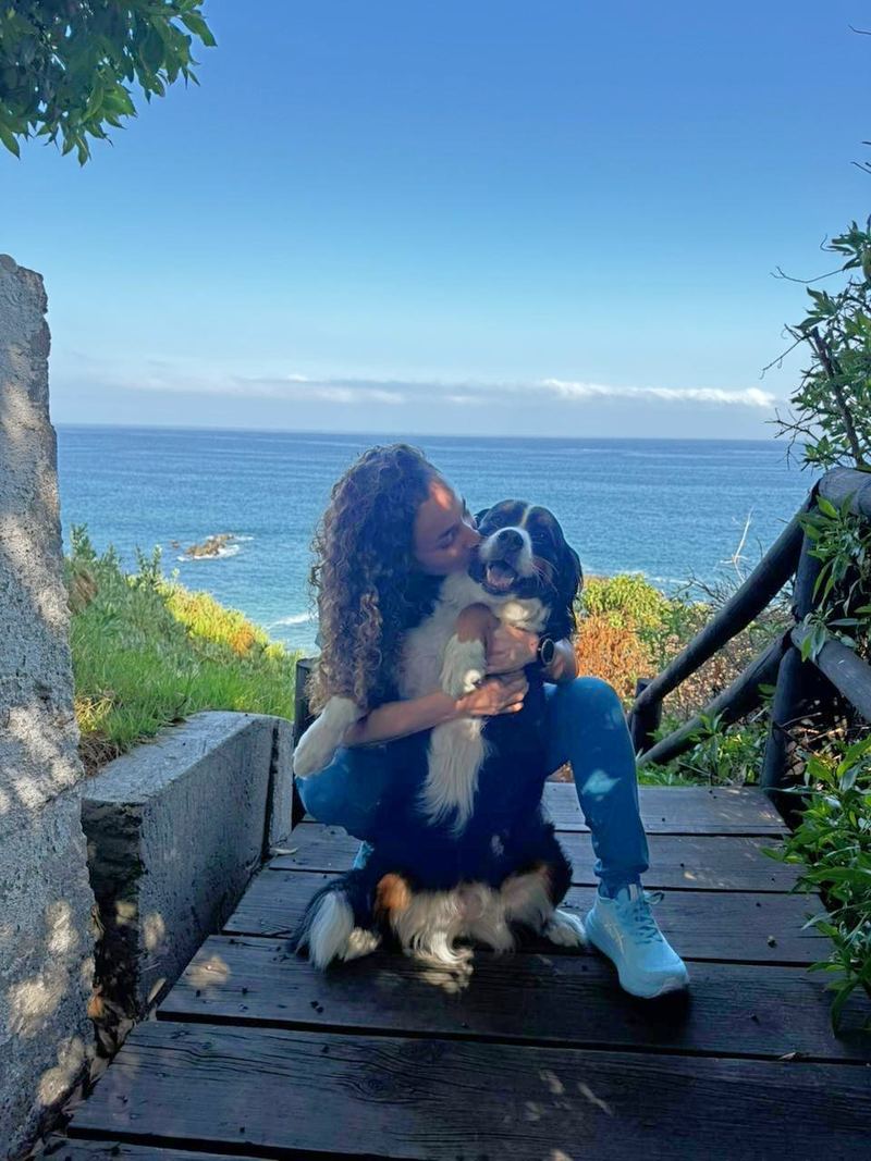 La Dra. Luna Fuentes sentada en una escalera de madera frente al mar, abrazando y dándole un beso a un perro tricolor